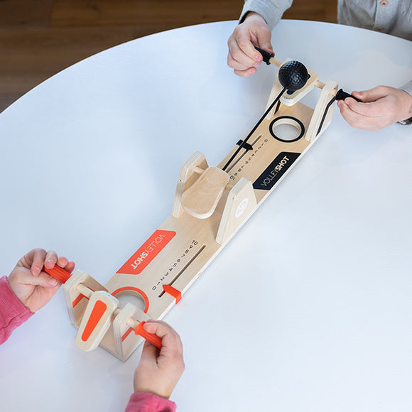 Two players engaging in a fun game of VolleyShot, a wooden game designed by Fat Brain. Each player holds a launcher, aiming to shoot a small ball through the center hole on a long, narrow wooden board. The game features colorful orange and black elements, and is set up on a white table, highlighting its interactive and competitive nature.