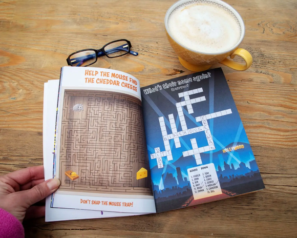 An open activity book titled 'Puzzled: The Frustratingly Futile Activity Book' showing a maze puzzle on the left page with a cartoon mouse and cheese, and a crossword puzzle on the right page featuring a blue city skyline background. A pair of glasses and a cup of coffee are placed beside the book on a wooden table.