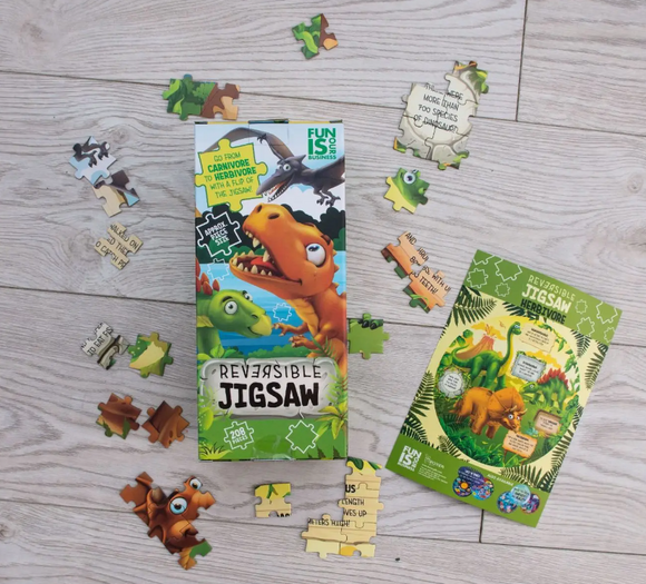 Colorful Dinosaurs Reversible Jigsaw puzzle by Boxer, featuring vibrant artwork of dinosaurs on the packaging. The image shows scattered puzzle pieces on a wooden floor, along with the packaging that includes descriptions of the features like carnivores and herbivores. Ideal for children, this jigsaw encourages learning and imaginative play as they piece together the fun dinosaur scenes.