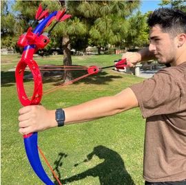 A young man in a brown t-shirt aims a red and blue toy bow called Ring Strike. The bow features red arrows and is being used in a grassy park setting. Trees and a picnic area are visible in the background.