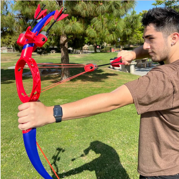 A young man in a brown t-shirt aims a red and blue toy bow called Ring Strike. The bow features red arrows and is being used in a grassy park setting. Trees and a picnic area are visible in the background.
