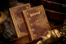 Set of Beowulf playing cards by Kings Wild, featuring intricate designs with a leather-like texture and gold detailing, displayed among decorative items.