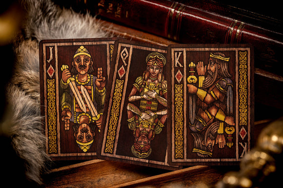 Image of Beowulf Playing Cards by Kings Wild, showcasing three cards featuring intricate illustrations of characters and symbols inspired by the Beowulf epic. The cards are displayed against a textured backdrop of fur and vintage wooden elements, giving a rustic feel to the overall presentation.