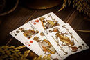 A close-up of Babylon Golden Wonders playing cards featuring intricate illustrations of kings and queens, showcasing rich details and colors, resting on a textured wooden surface surrounded by natural elements.