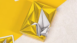 1000 Cranes V3 Playing Cards by Riffle Shuffle. The card box features a geometric design with yellow and gray tones, showcasing a modern and artistic look.