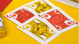A close-up view of 1000 Cranes V3 Playing Cards featuring intricately designed cards in vibrant red and yellow colors. The cards showcase geometric patterns and symbols, placed on a bright yellow background alongside a silver box.