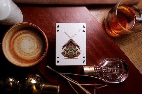 A beautifully designed playing card from Rattler Gorge titled 'Ace of Spades' featuring intricate graphics on a wooden surface. Surrounding the card are a handcrafted bowl, a bottle of whiskey, and decorative items, creating a cozy atmosphere perfect for game nights.