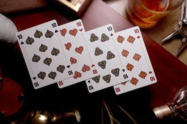 Rattler Gorge Playing Cards (Desert Dust) displayed on a wooden surface with various vintage items in the background. The cards show intricate designs with hearts and spades symbols, featuring contrasting colors of red and black. Ideal for card games and collectors alike.