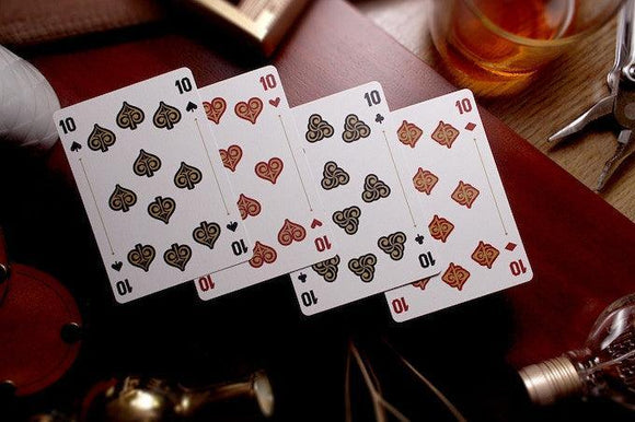 Rattler Gorge Playing Cards (Desert Dust) displayed on a wooden surface with various vintage items in the background. The cards show intricate designs with hearts and spades symbols, featuring contrasting colors of red and black. Ideal for card games and collectors alike.