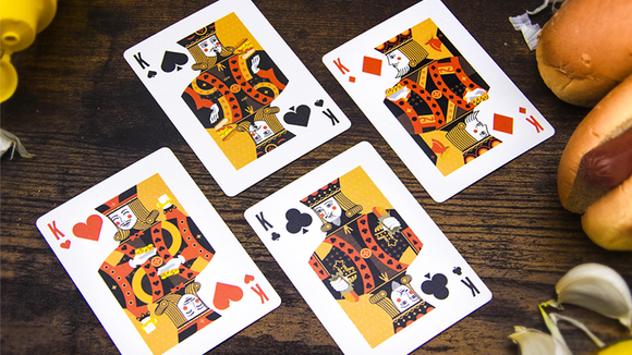 Four playing cards featuring unique designs with a hot dog theme. The cards display Kings from different suits: Spades, Hearts, Diamonds, and Clubs, each adorned with vibrant colors and playful graphics. They're arranged on a wooden surface next to some real hot dogs, creating a fun and lighthearted gaming atmosphere.