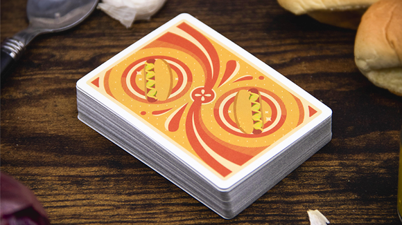 A deck of Hot Dog Playing Cards by Fast Food Playing Cards displayed on a wooden surface. The card back features a vibrant design with two illustrated hot dogs on a swirl pattern, surrounded by playful decorative elements. Nearby are some hot dog buns and a spoon, enhancing the fun food theme.