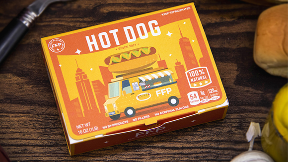 A colorful box of Hot Dog Playing Cards by Fast Food Playing Cards. The box features an illustration of a food truck with a hot dog on top, set against a city skyline backdrop. The product highlights its 100% natural ingredients and playful design, making it an engaging addition to any game night.
