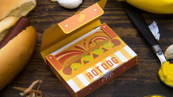 A brightly colored box of Hot Dog Playing Cards featuring an illustration of a hot dog with mustard and toppings. The box is partially open, revealing the card design inside, and is surrounded by a hot dog bun, mustard, and garlic, set against a wooden surface.