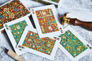 An array of Bloodlines Emerald Playing Cards displayed on a textured surface. The cards feature vibrant, intricate illustrations with a blend of red, green, and yellow colors. A partially visible decorative box and a wooden stamp are also present, adding to the visual composition.