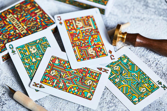 An array of Bloodlines Emerald Playing Cards displayed on a textured surface. The cards feature vibrant, intricate illustrations with a blend of red, green, and yellow colors. A partially visible decorative box and a wooden stamp are also present, adding to the visual composition.