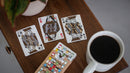 A set of Roasters Coffee Shop playing cards featuring artistic designs. Three cards are shown: the Jack of Spades, the Queen of Hearts, and the King of Spades, placed on a wooden table alongside a coffee cup and a colorful card holder.