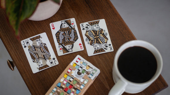 A set of Roasters Coffee Shop playing cards featuring artistic designs. Three cards are shown: the Jack of Spades, the Queen of Hearts, and the King of Spades, placed on a wooden table alongside a coffee cup and a colorful card holder.