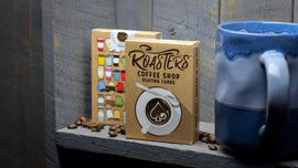Roasters Coffee Shop Playing Cards by US Playing Card Co. The deck features a playful coffee theme with colorful cup designs on the packaging, perfect for coffee lovers and card game enthusiasts. The cards are displayed next to a blue ceramic mug and scattered coffee beans, set against a rustic wooden background.