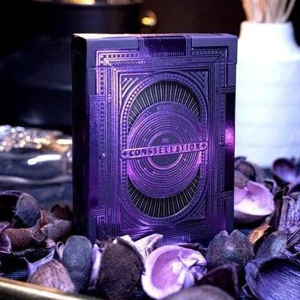 The Constellation Majestic Playing Cards by Deckidea, elegantly presented in a decorative setting with purple and natural textures. The packaging features intricate designs and shimmering accents, showcasing its premium quality ideal for card enthusiasts.