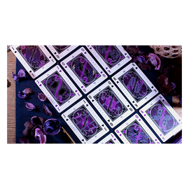 The Constellation Majestic Playing Cards on a dark blue surface, featuring intricate designs in purple and black. The cards are spread out, showcasing unique illustrations and heavenly motifs. Surrounding the cards are dried purple petals, adding a touch of elegance to the arrangement.