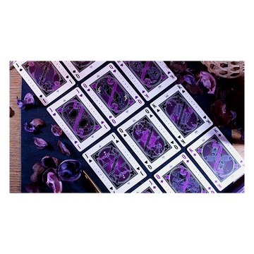 The Constellation Majestic Playing Cards on a dark blue surface, featuring intricate designs in purple and black. The cards are spread out, showcasing unique illustrations and heavenly motifs. Surrounding the cards are dried purple petals, adding a touch of elegance to the arrangement.