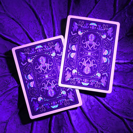 A close-up view of Bioluminescent Playing Cards featuring vibrant purple and blue designs with aquatic imagery. The cards are set against a textured purple background, highlighting their glowing effects in low light.