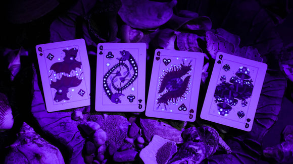 A set of bioluminescent playing cards displayed on a bed of leaves and stones, featuring illustrations of bats, caterpillars, and mushrooms, glowing under purple light. Each card showcases intricate designs that come alive in the dark, perfect for both gameplay and collectors.