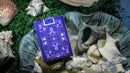A box of Bioluminescent Playing Cards by Penguin Magic, displayed on a bed of seashells and moss. The elegantly designed box features vibrant purple hues with intricate illustrations of octopuses and decorative patterns, showcasing its unique and whimsical theme.
