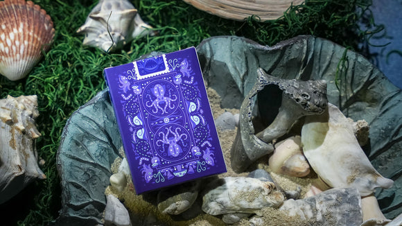 A box of Bioluminescent Playing Cards by Penguin Magic, displayed on a bed of seashells and moss. The elegantly designed box features vibrant purple hues with intricate illustrations of octopuses and decorative patterns, showcasing its unique and whimsical theme.