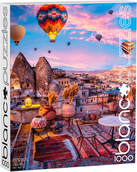 Blanc Puzzles 1000-piece jigsaw featuring a stunning scene of colorful hot air balloons floating above the unique rock formations of Cappadocia at sunset. The image includes traditional pottery and cozy furniture set against an enchanting landscape, perfect for puzzle lovers and travel enthusiasts.