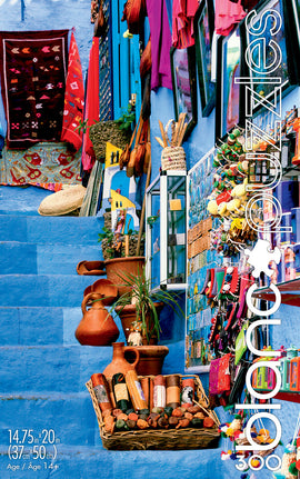A vibrant jigsaw puzzle featuring a scenic view of the Blue City in Morocco, showcasing colorful crafts, textiles, and pottery. The image captures intricate details of a charming street adorned with traditional Moroccan decor, inviting puzzlers to explore this delightful landscape piece by piece. The puzzle measures 14.75 in x 20 in (37 cm x 50 cm) and is designed for ages 14 and above.