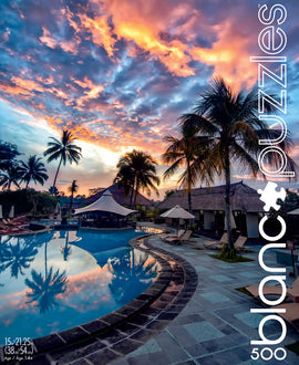 A vibrant jigsaw puzzle titled 'Bali Sunset' by Blanc Puzzles, featuring a stunning sunset over a serene pool surrounded by luxurious villas, palm trees, and a colorful sky, perfect for relaxation and leisure. The puzzle consists of 500 pieces, measuring 15x21.25 inches, designed for ages 14 and up.