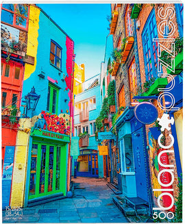 A vibrant jigsaw puzzle image depicting a colorful narrow street in London, featuring brightly painted buildings in hues of blue, yellow, and pink. A shop named 'Hair by Fairy' is prominently displayed, along with hanging plants and outdoor lighting. The scene captures the charm of urban life with colorful façades and a clear blue sky in the background.