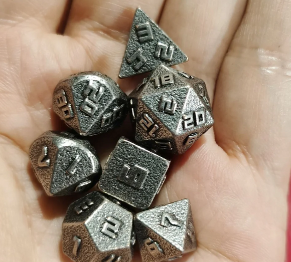 A hand holding a collection of seven 10MM ancient silver RPG dice in various shapes including a tetrahedron, cube, and dodecahedron. The dice are engraved with numbers and have a textured silver finish, perfect for tabletop gaming.
