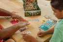 An engaging tabletop scene featuring the board game 'Oh No, Volcano!' by Buffalo Games. Two players interact with colorful game cards and tokens spread out on a wooden table, while the game box showcases a vibrant illustration of explorers in a tropical setting. One player is visibly strategizing with hands pointing at different cards to make decisions during gameplay.