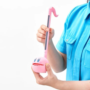 A person holding an Otamatone Kirby, a whimsical musical instrument shaped like a cartoon character. The pink body features a smiling face and is being played with a curved, pink musical wand. The setting has a bright background, showcasing the playful design of this unique item.