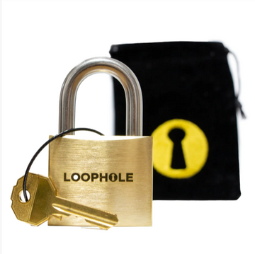 The Loophole mechanical puzzle features a shiny brass padlock with a distinctive logo engraved on the front. Attached to the padlock is a key, which is partially visible. The puzzle is displayed next to a black velvet pouch that has a yellow keyhole design, suggesting an element of mystery and challenge in unlocking the padlock.