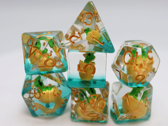 A close-up image of Green Dragon Hatchlings RPG Dice by Foam Brain. The dice are translucent with an aqua blue tint and embedded yellow dragon hatchling figures. Each die features gold numbers that contrast beautifully with the colorful design, perfect for tabletop RPG enthusiasts.