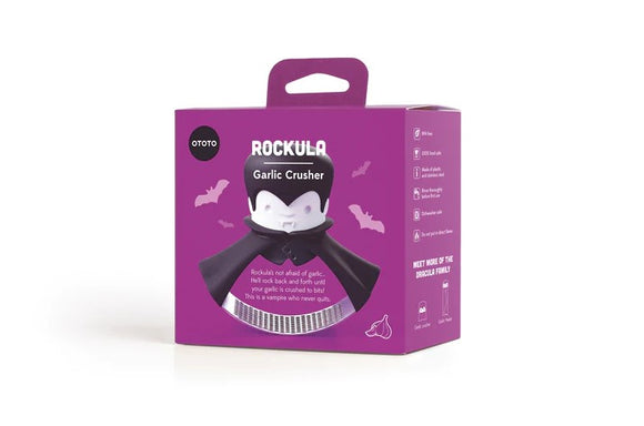 The Rockula Garlic Press by OToto is packaged in a vibrant purple box featuring a whimsical vampire character in a cape. The character's playful design comes with bat motifs surrounding it, inviting users to crush garlic with ease. The packaging highlights the fun and practicality of the garlic press, making it a unique kitchen tool.
