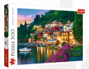 Trefl 500-piece jigsaw puzzle featuring a picturesque view of Lake Como, Italy. The image showcases colorful houses along a serene waterfront, surrounded by lush greenery and vibrant flowers, under a twilight sky. Perfect for puzzle enthusiasts and Italy lovers.