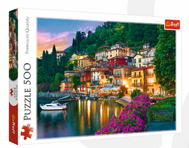 Trefl 500-piece jigsaw puzzle featuring a picturesque view of Lake Como, Italy. The image showcases colorful houses along a serene waterfront, surrounded by lush greenery and vibrant flowers, under a twilight sky. Perfect for puzzle enthusiasts and Italy lovers.