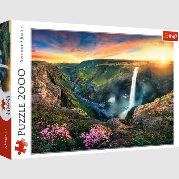 Trefl jigsaw puzzle featuring the breathtaking Háifoss Waterfall in Iceland. The scene captures a stunning sunrise over a lush, green valley with vibrant pink flowers in the foreground. The waterfall cascades dramatically down rocky cliffs, with a serene river winding through the landscape, creating a picturesque view that's perfect for puzzle lovers.