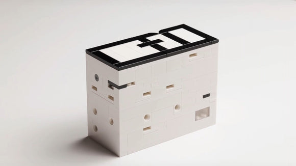 The aMazing PuzzleBox by QuizBrix is a mechanical puzzle featuring a sleek, white design with black accents. It has various openings and slots, encouraging problem-solving and creativity as users figure out how to unlock its secrets.