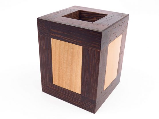 The Identical Twins mechanical puzzle by Pelikan Puzzles features a beautifully crafted wooden cube with a square opening at the top. The puzzle is made of rich, dark wood with lighter wood insets, showcasing intricate craftsmanship and a sophisticated design. Ideal for puzzle enthusiasts looking for a challenging and aesthetic addition to their collection.