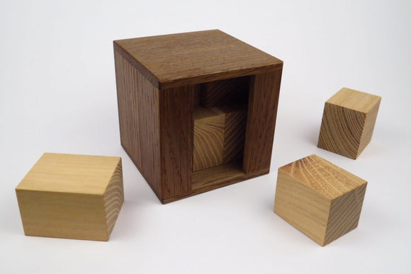 The Euklid mechanical puzzle by Pelikan Puzzles features a wooden cube with a sliding mechanism. Four cubes are displayed, two light-colored and two dark-colored, each with unique wood grain patterns. The design encourages problem-solving and dexterity as players maneuver the cubes into the main box.
