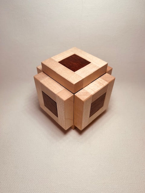 A beautifully crafted wooden mechanical puzzle called Coaxial, made by Pelikan Puzzles. The puzzle features a cube shape with distinct natural wood patterns, showcasing both light and dark wood tones. Each side has a unique design that invites interaction and problem-solving, making it a perfect gift for puzzle enthusiasts.