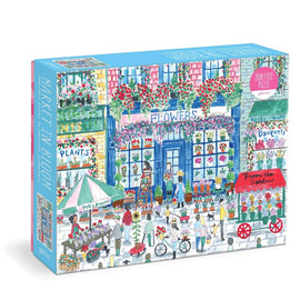 Michael Storrings Market in Bloom jigsaw puzzle by Galison, featuring a vibrant scene of a flower market with colorful stalls, people buying flowers, and a charming street lined with plants and bouquets. This 2000-piece puzzle captures the essence of a bustling open-air market, perfect for puzzle enthusiasts.