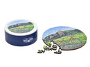 The Boulder Picnic jigsaw puzzle from Liberty Puzzles features a picturesque scene of a bicycle set against a vibrant landscape with mountains and greenery. The round puzzle is partially assembled, with colorful, intricately shaped wooden pieces scattered around a decorative tin container emblazoned with the Liberty Puzzles logo.
