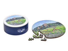 The Boulder Picnic jigsaw puzzle from Liberty Puzzles features a picturesque scene of a bicycle set against a vibrant landscape with mountains and greenery. The round puzzle is partially assembled, with colorful, intricately shaped wooden pieces scattered around a decorative tin container emblazoned with the Liberty Puzzles logo.
