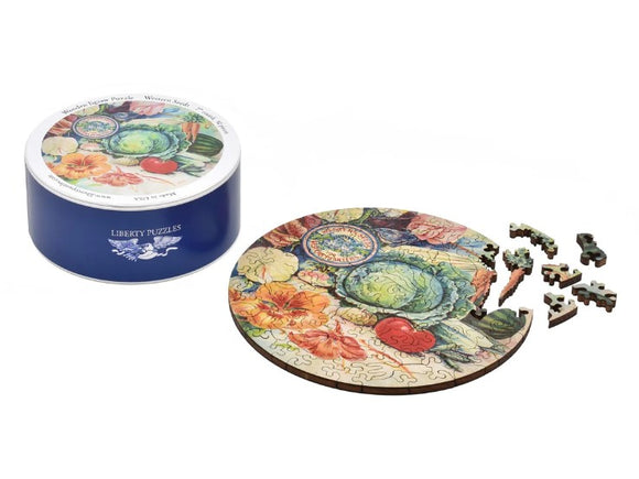 A round jigsaw puzzle titled 'Western Seeds' by Liberty Puzzles, featuring vibrant floral and vegetable illustrations. The puzzle is partially assembled, with several pieces scattered nearby. The puzzle comes in a decorative tin with a colorful lid showcasing the same artwork.